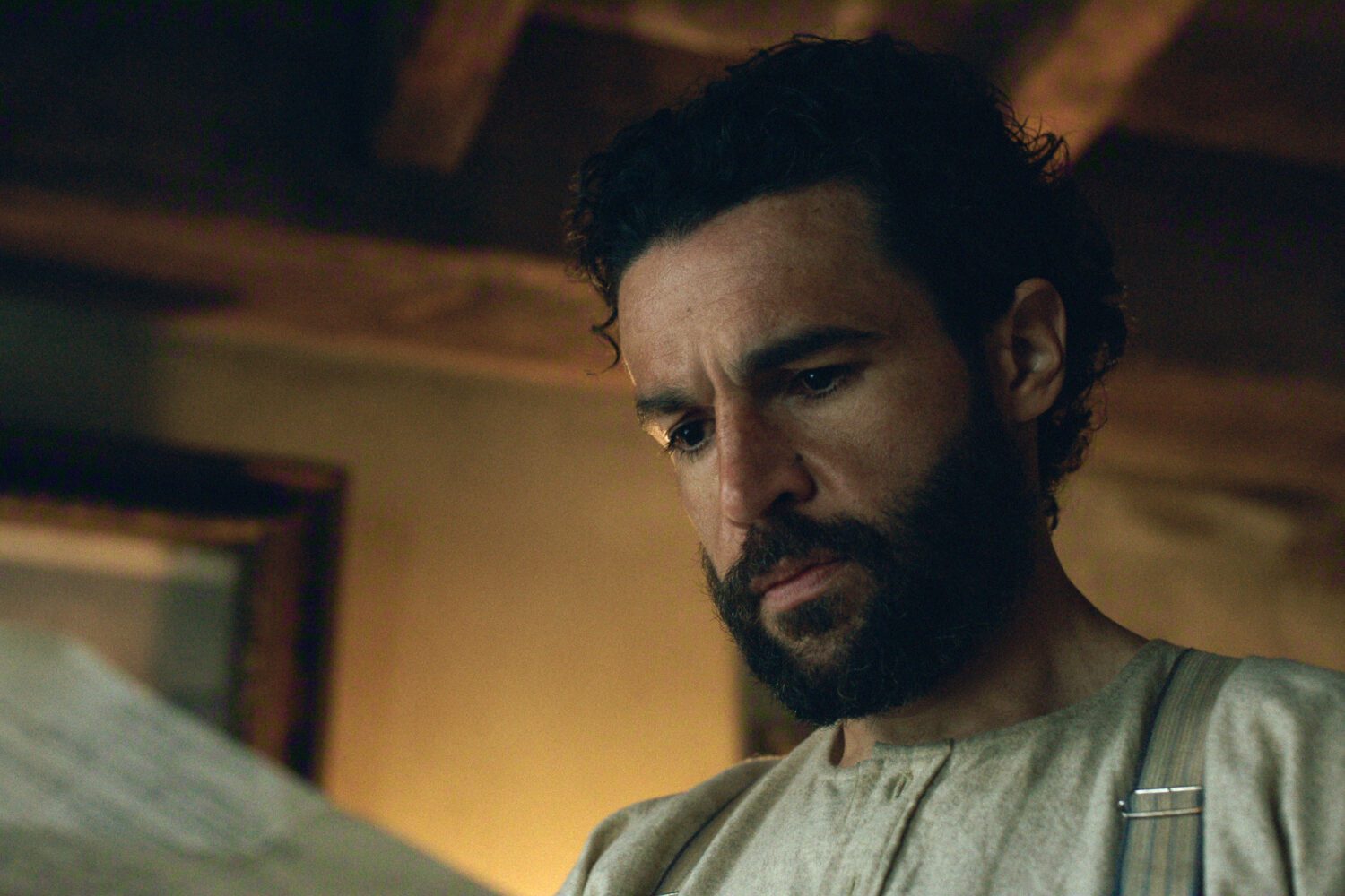 "A man with a beard wearing a simple garment looks thoughtfully at a piece of paper indoors, in a dimly lit, rustic room with wooden beams and an old framed picture in the background."